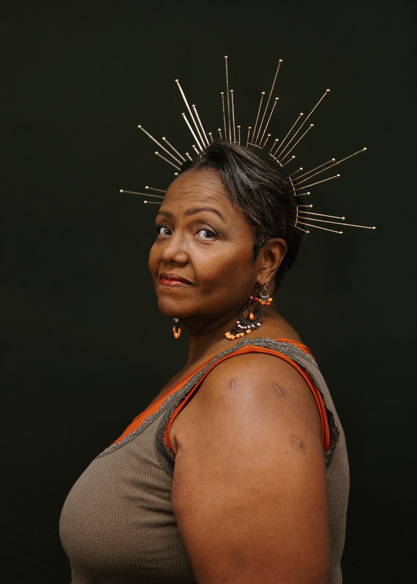 Woman with short gray hair wears a gold sunburst crown and orange beaded earrings, looking over her shoulder against a dark background.