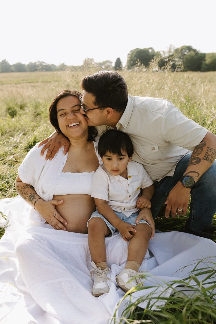 Family in white clothing shares tender moment during outdoor maternity photoshoot in sunny field.