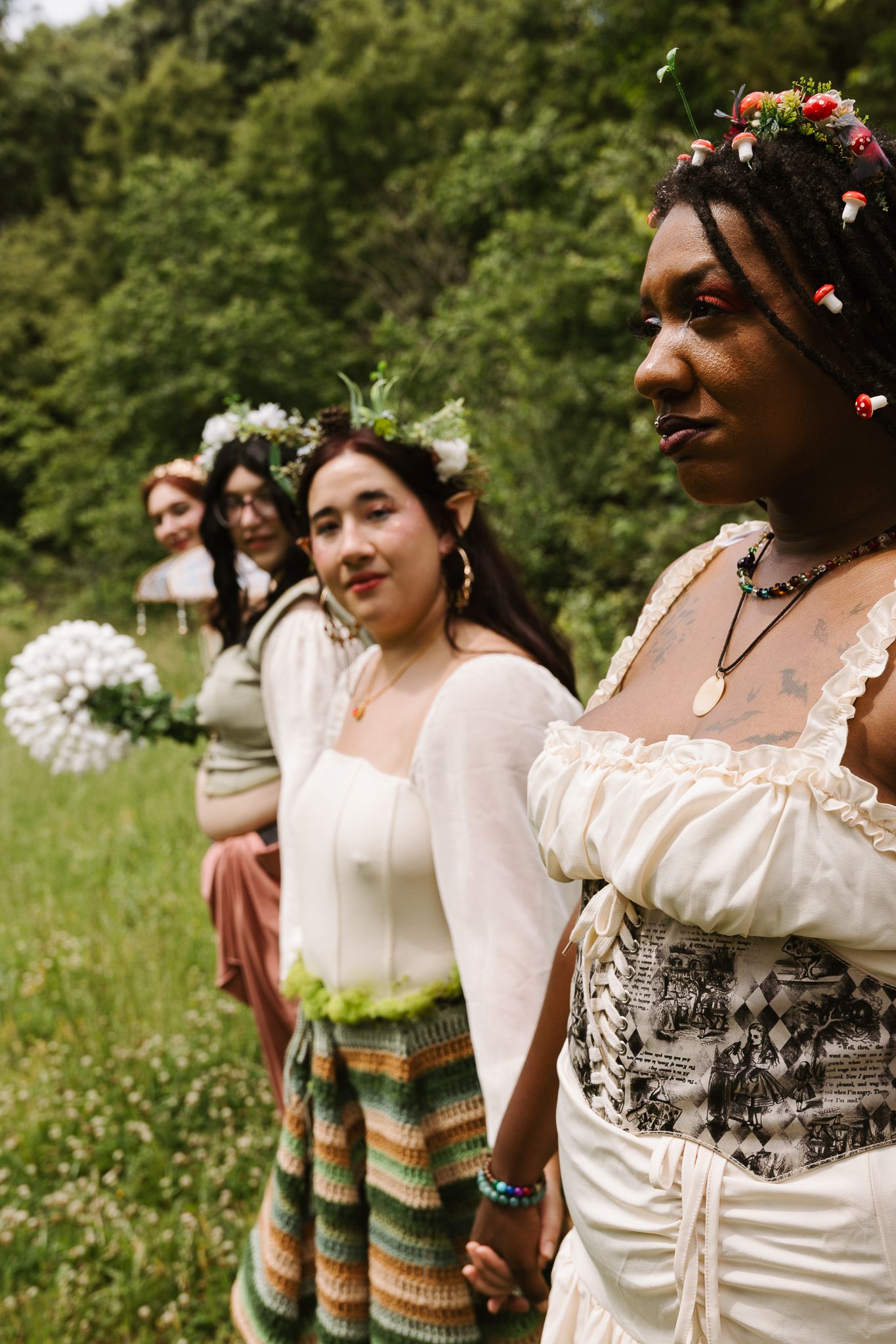 Four diverse women in white boho dresses and floral crowns stand in a lush green meadow during an outdoor celebration.
