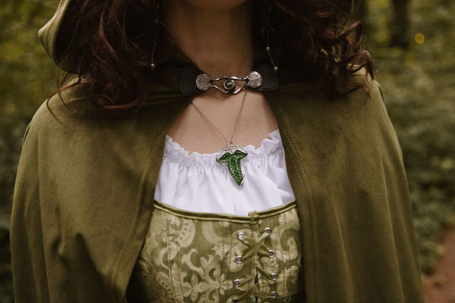 Woman in medieval costume with green leaf pendant necklace and olive cloak in forest setting.