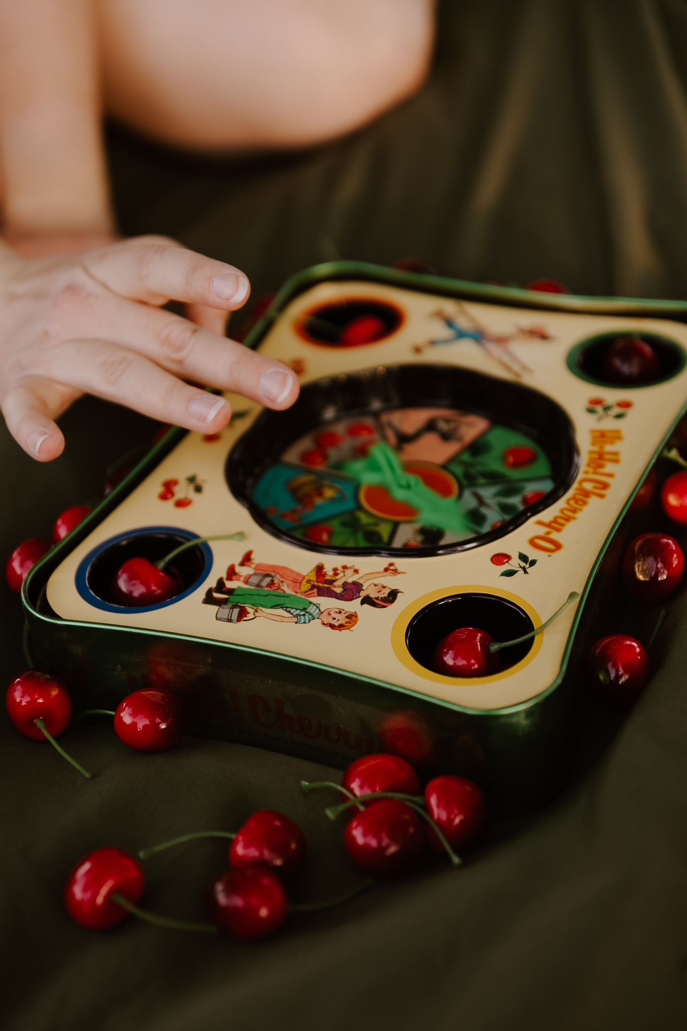 A child's hand reaches toward a vintage cherry-themed tin game board surrounded by fresh red cherries on green fabric.