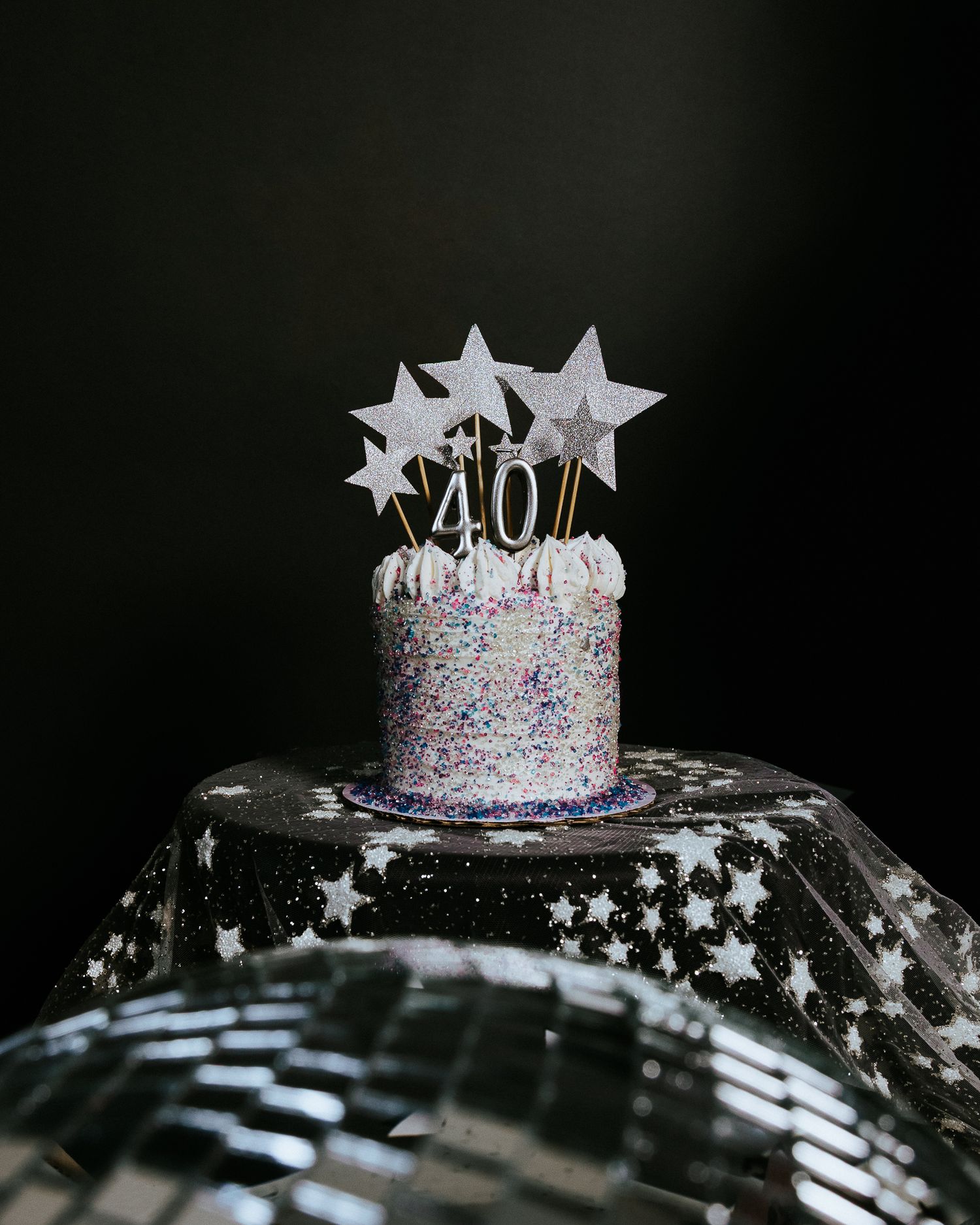 A glittery 40th birthday cake topped with silver stars sits on a disco ball against a dark background.