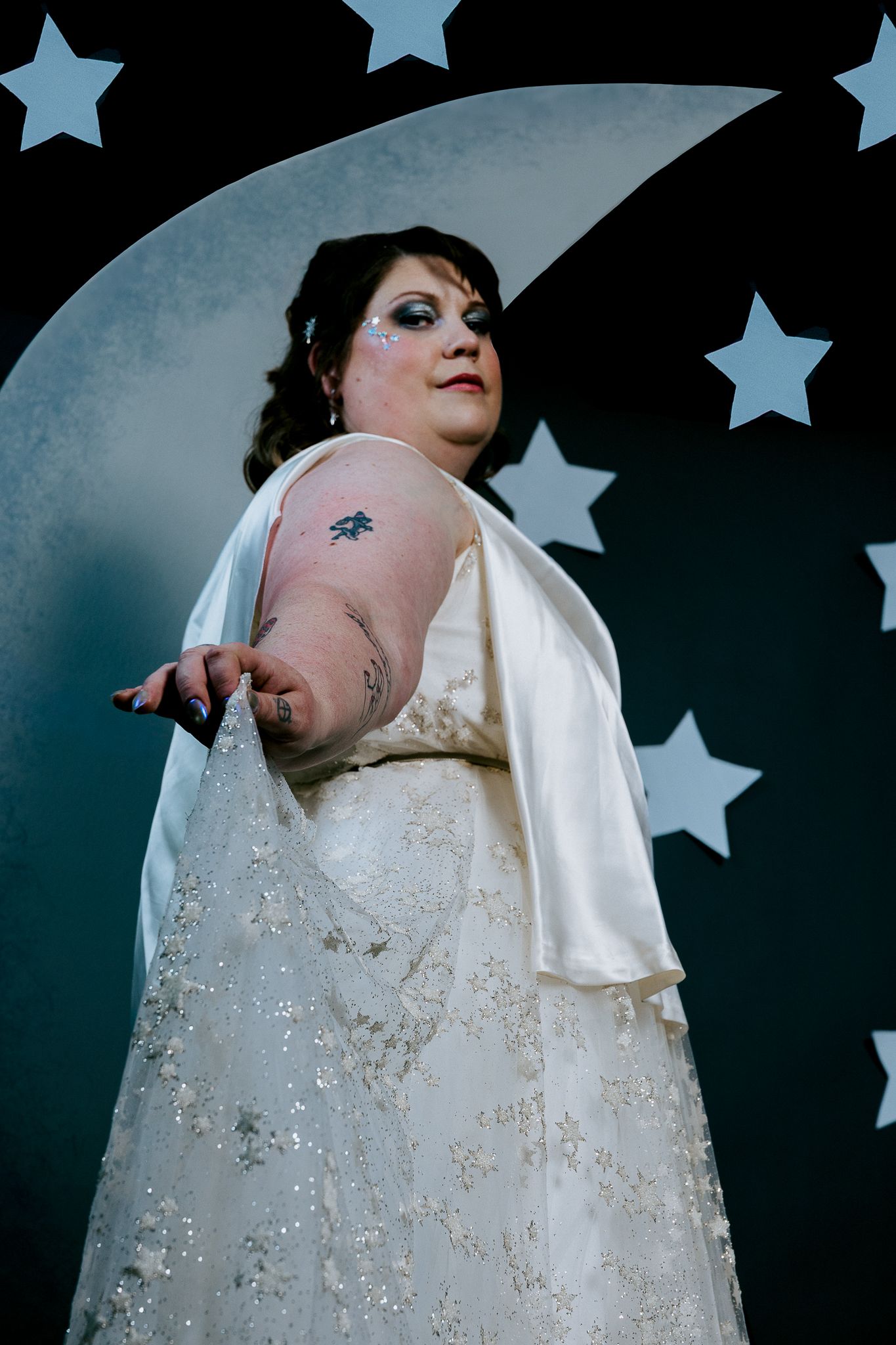 Plus-size woman in white sequin gown poses before moon and stars backdrop with dramatic makeup and arm tattoo.