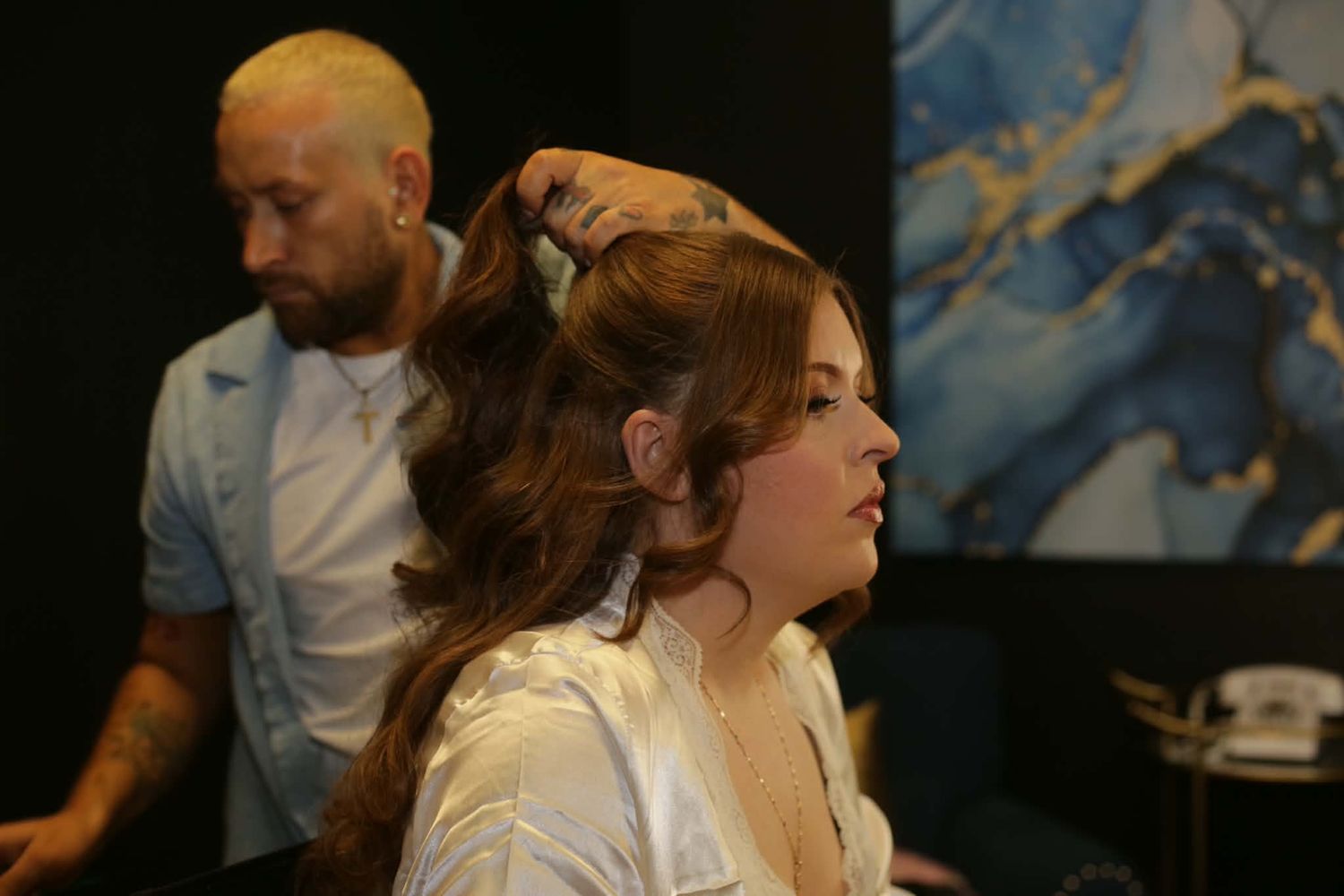 A male hairstylist with blonde hair styles a young womans long wavy auburn hair in a modern salon.