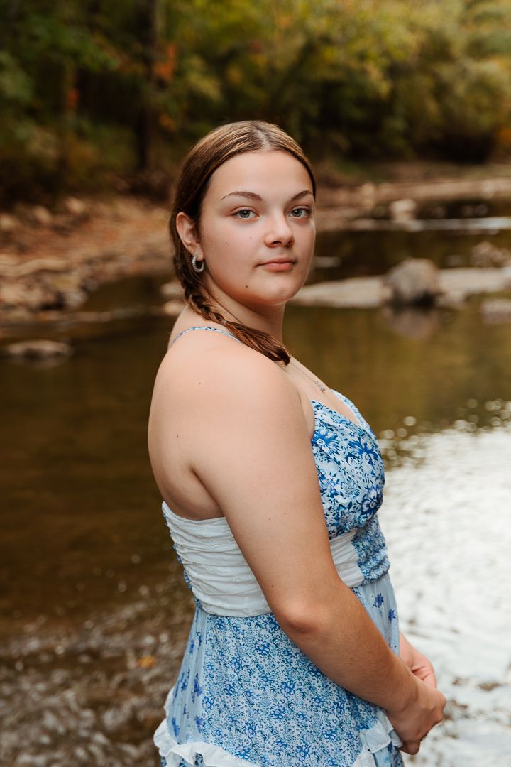 Portrait in a blue floral dress poses by a tranquil stream in an autumn woodland setting.