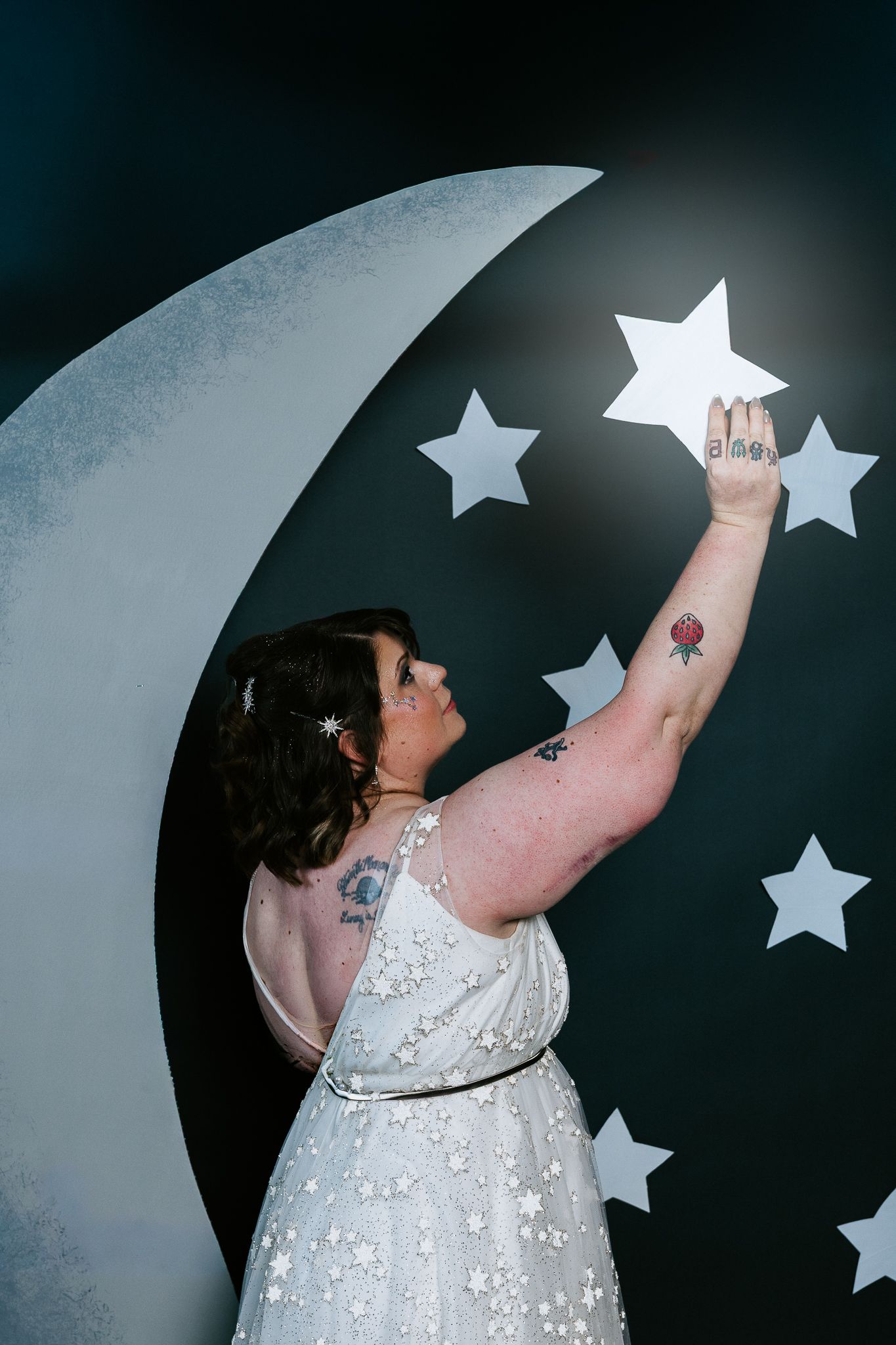 A tattooed woman in a white floral dress reaches up to place a star on a decorative moon backdrop.