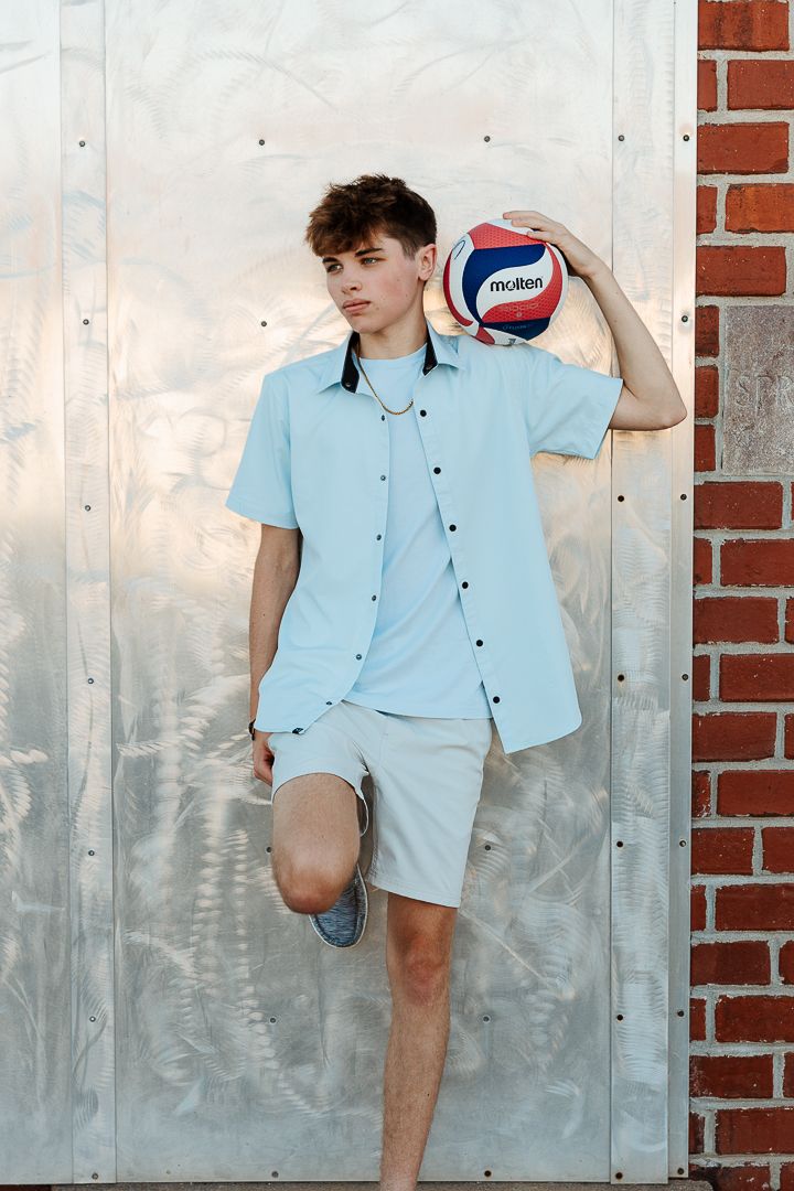 Young model in light blue button-down shirt poses against metallic wall with brick accent.