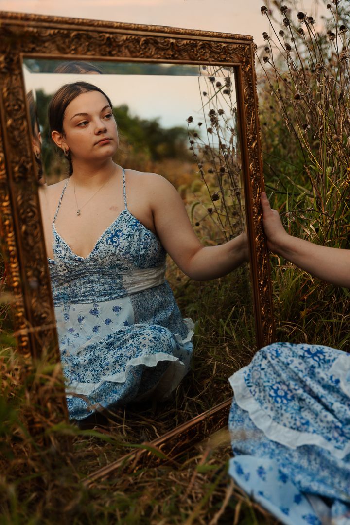A vintage-framed mirror reflects a blue dress against wild grass and flowers in golden hour light.