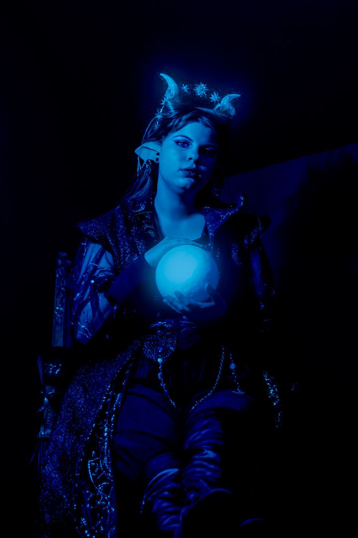 A fantasy elf queen in ornate armor holds a glowing blue orb in dramatic dark blue lighting.