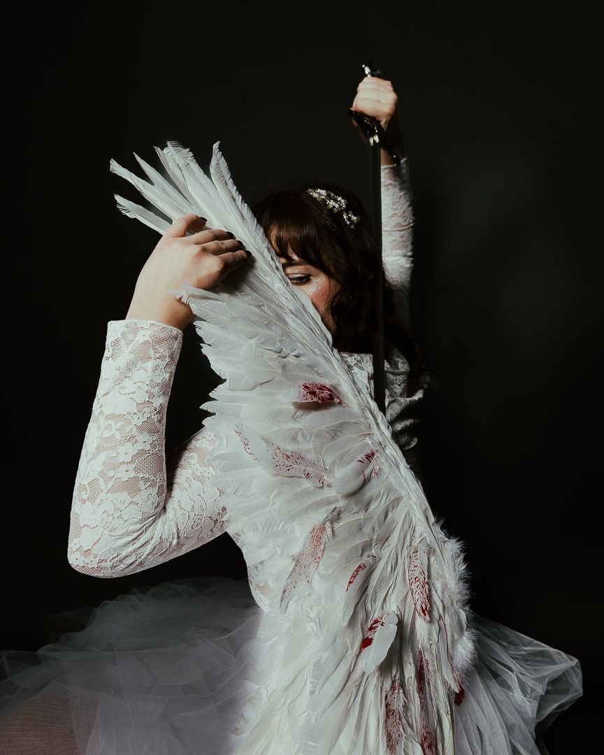 A woman in a blood-stained white lace dress holds feathered wings against a dark background.
