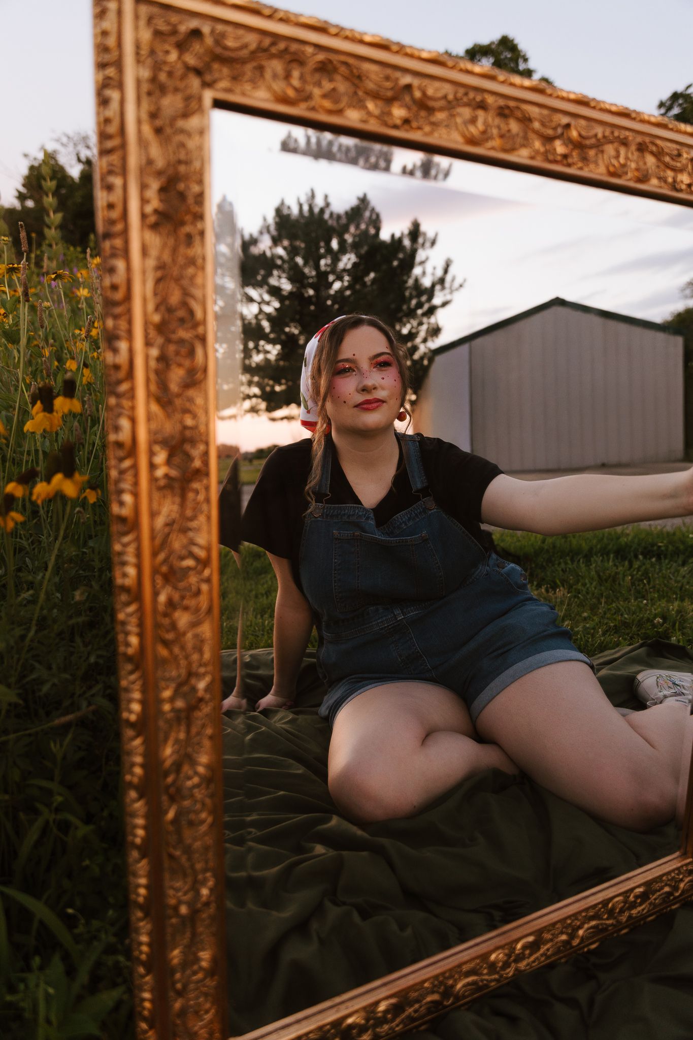 Young woman in denim overalls sits outdoors framed by an ornate gold picture frame at golden hour.