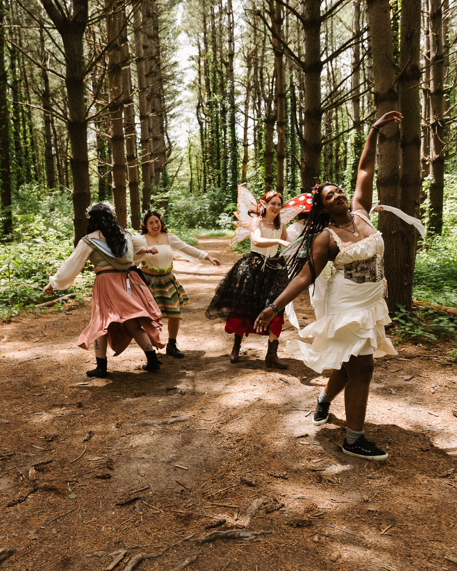 Five women in colorful fairy-tale costumes dancing joyfully in a sunlit forest clearing among tall trees.