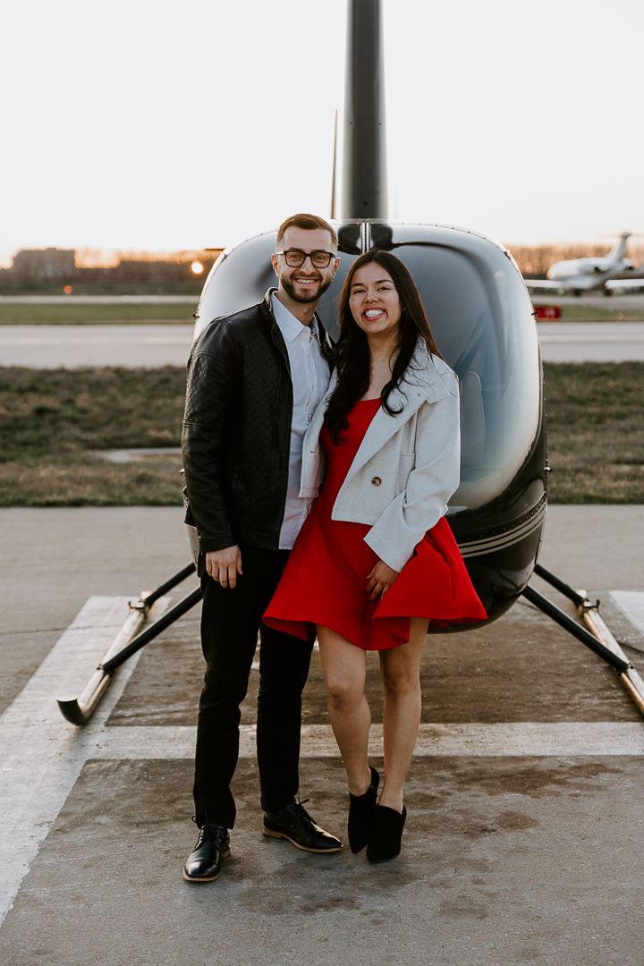 A couple in formal wear poses playfully in front of a small aircraft during sunset.