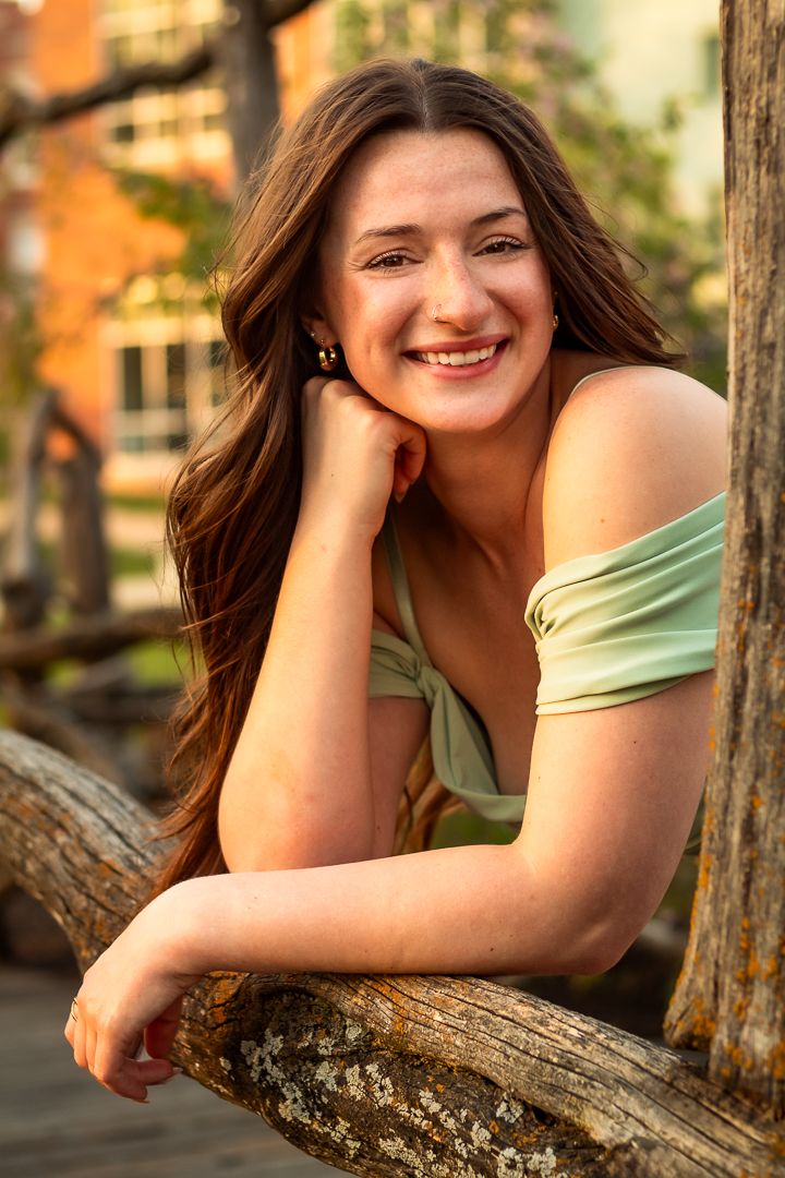 Portrait of someone outdoors in a green off-shoulder top leaning on rustic wood with a warm sunset glow in the background.