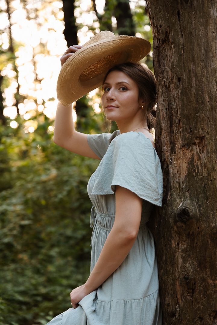Person in vintage-style dress gazes upward while standing against a large tree trunk in dappled forest light.