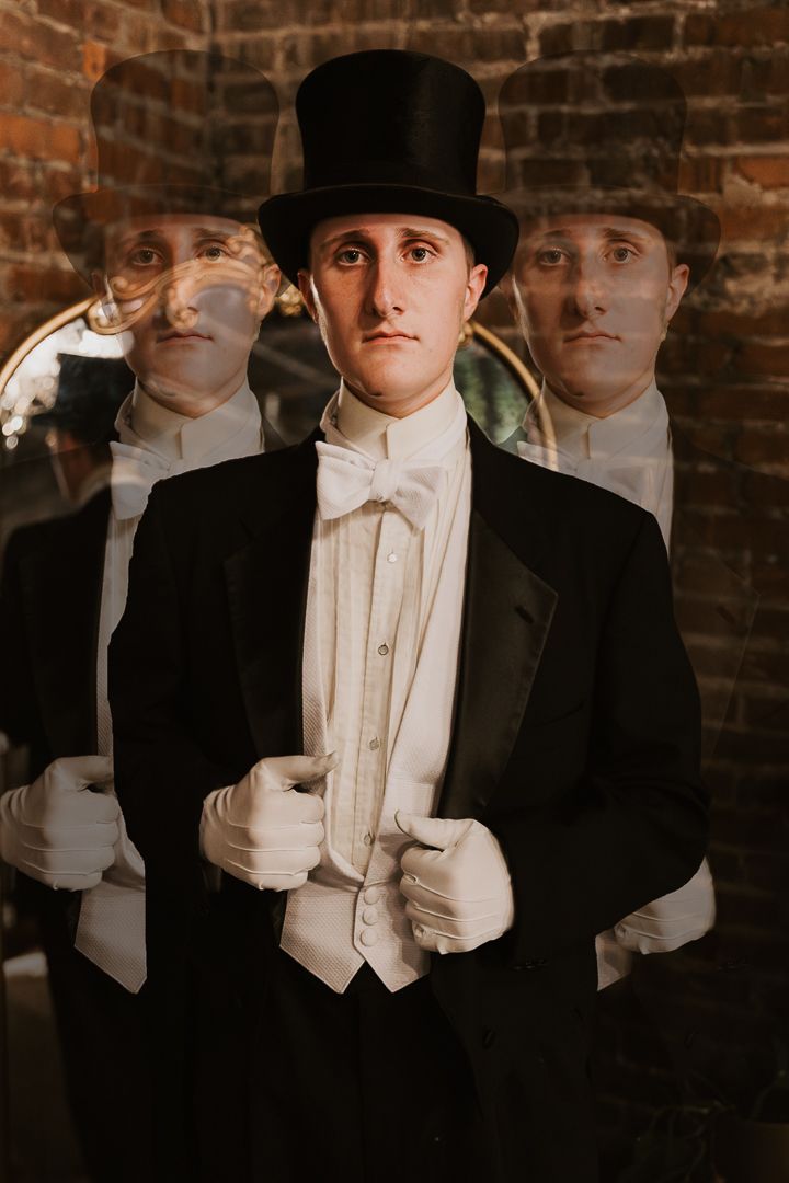 A person in formal evening attire and white gloves stands against a brick wall in a vintage-style portrait.