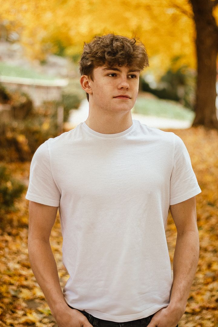 Someone in a white t-shirt poses against vibrant autumn foliage with golden yellow leaves in the background.