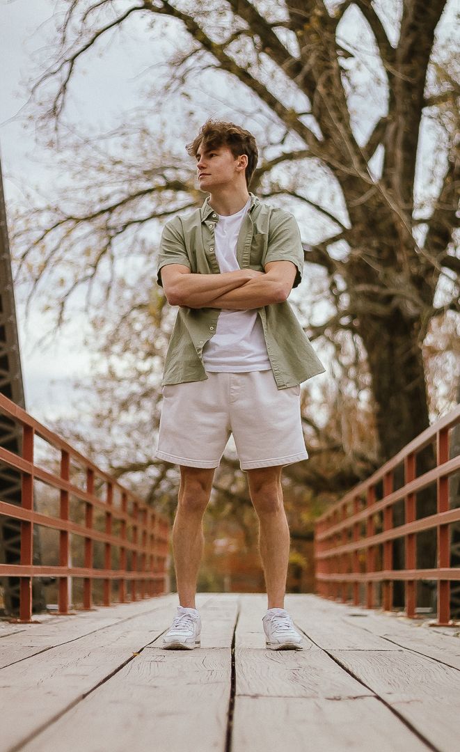 Low angle shots of someone in khaki shorts and white sneakers standing on a wooden boardwalk surrounded by autumn trees.