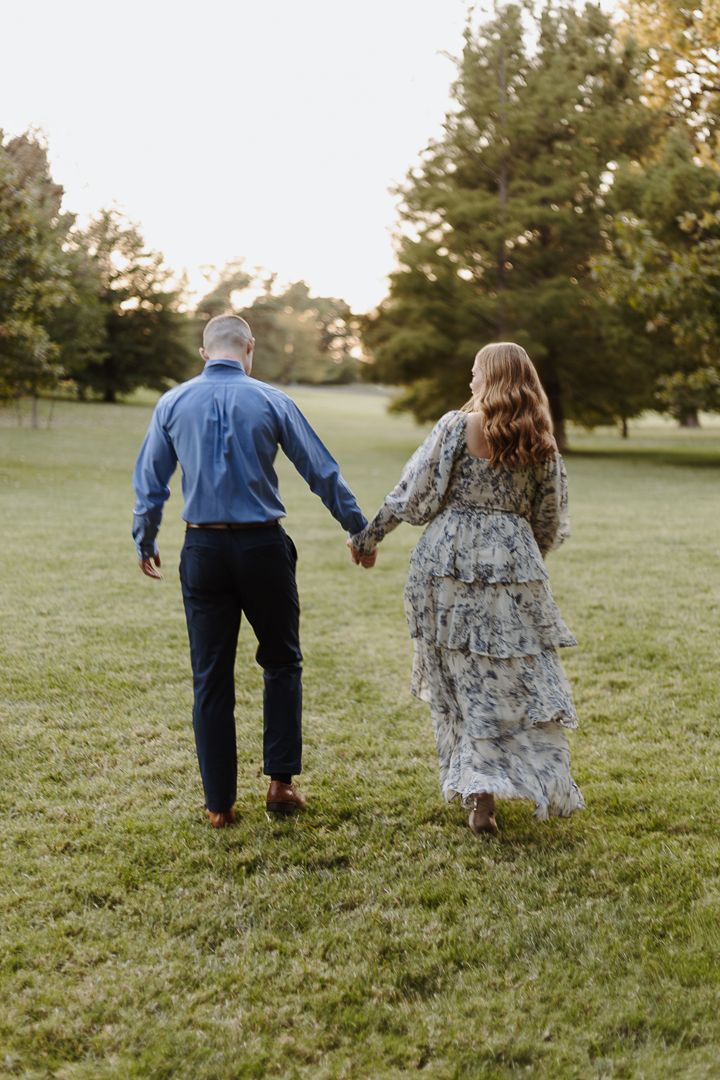 A couple walks hand in hand through a grassy field at sunset, with trees in the background.