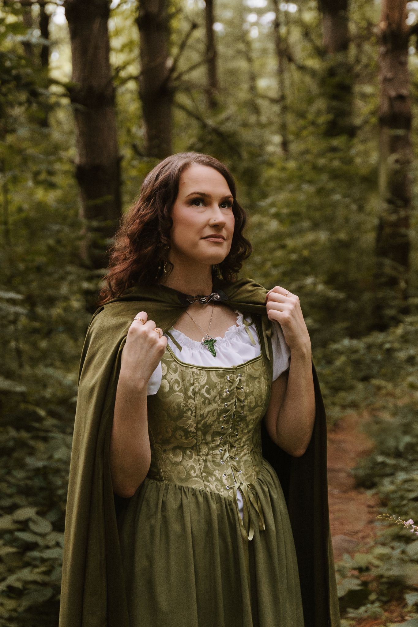 Woman in green medieval gown and cape poses in forest, Lord of the Rings inspired fantasy costume.
