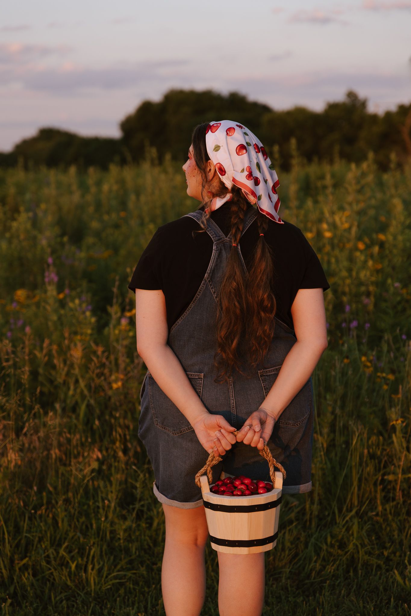 Woman in denim overalls holding a berry basket in a wildflower field at sunset, wearing a polka dot headscarf.