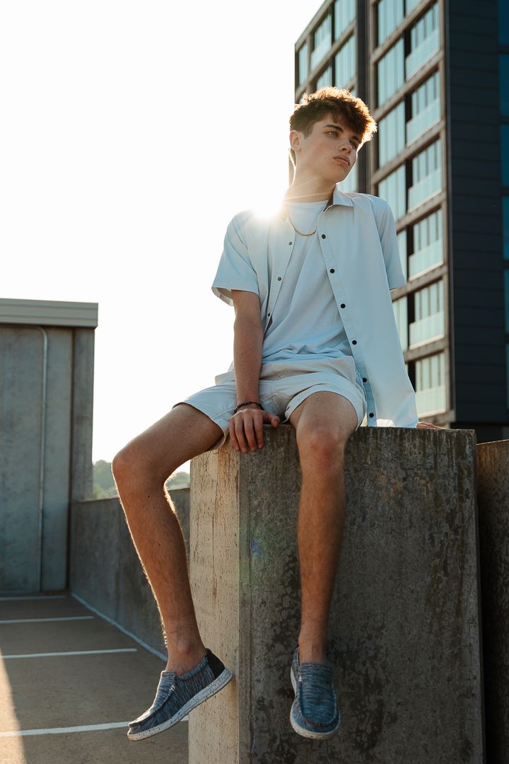 Person in light blue summer outfit and sneakers sits on concrete ledge during golden hour with modern building in background.