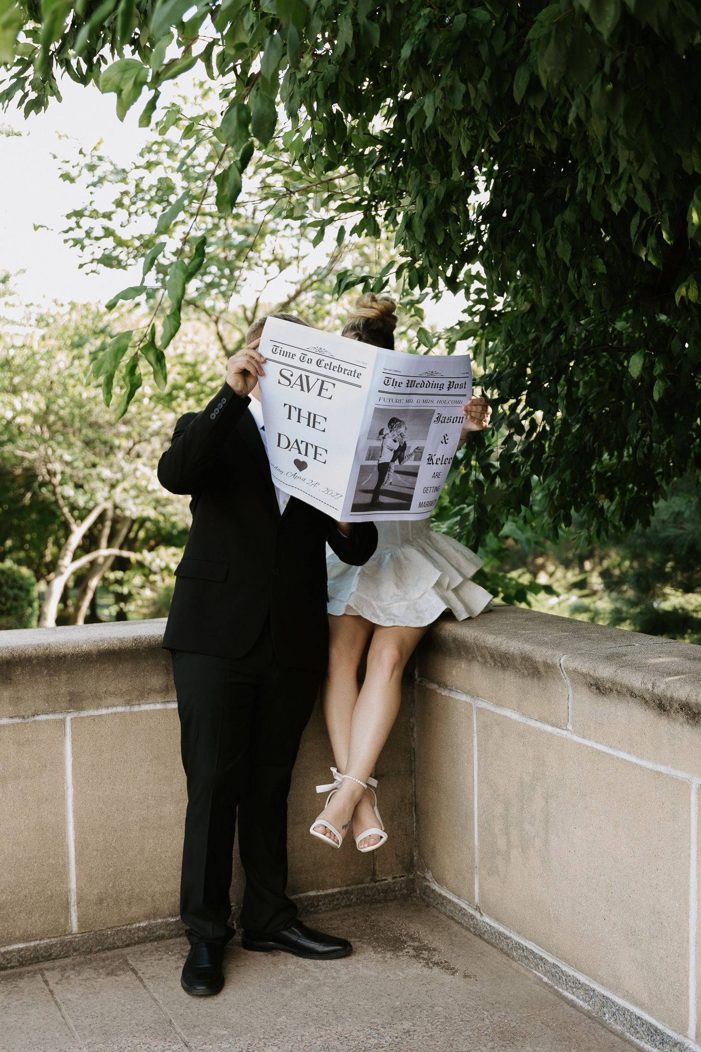 Couple hiding behind a Save the Date newspaper announcement, dressed in wedding attire, surrounded by lush green trees.