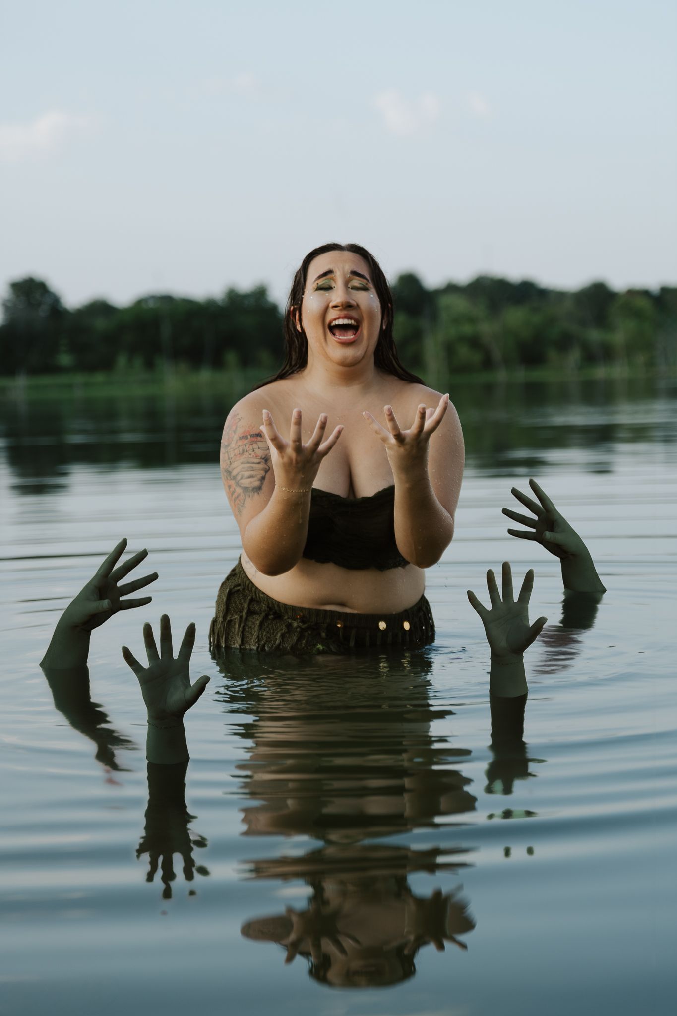 Woman standing in a lake screaming with green hands emerging from the water around her in a spooky scene.