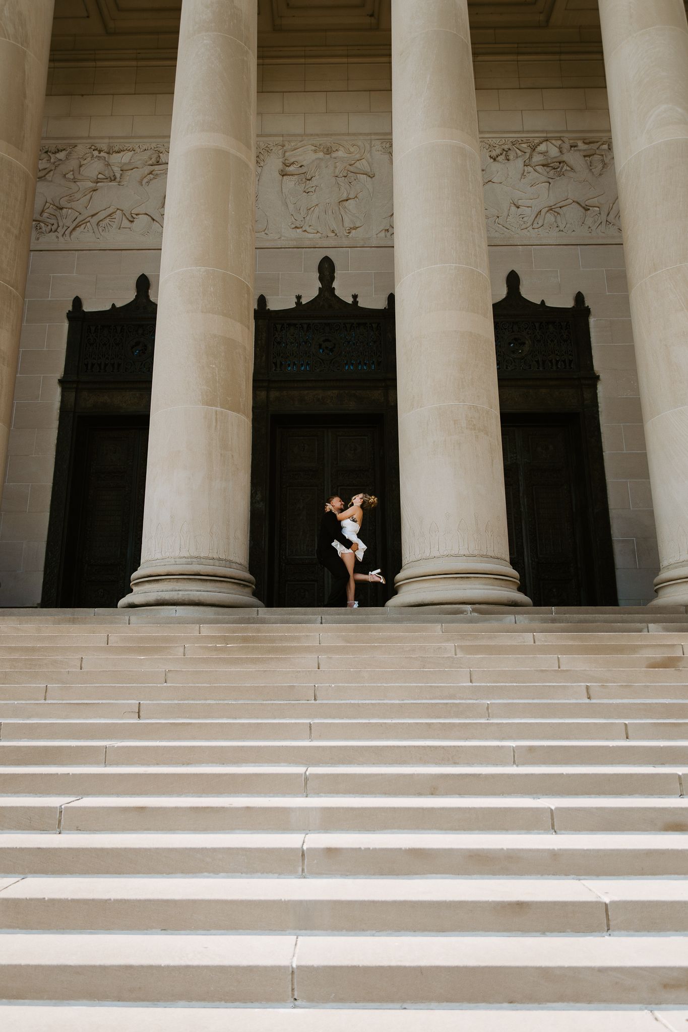 Couple embracing on grand stone steps of a neoclassical building with tall columns and ornate friezes.