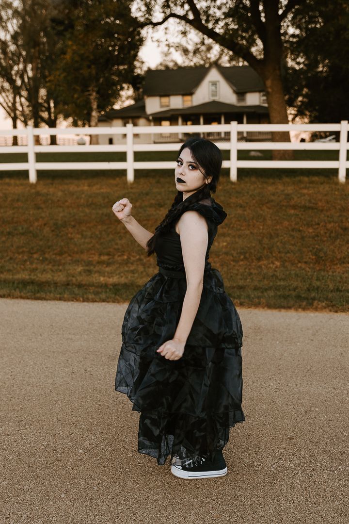 A person in a flowing black dress sits on pavement near a white picket fence at sunset.