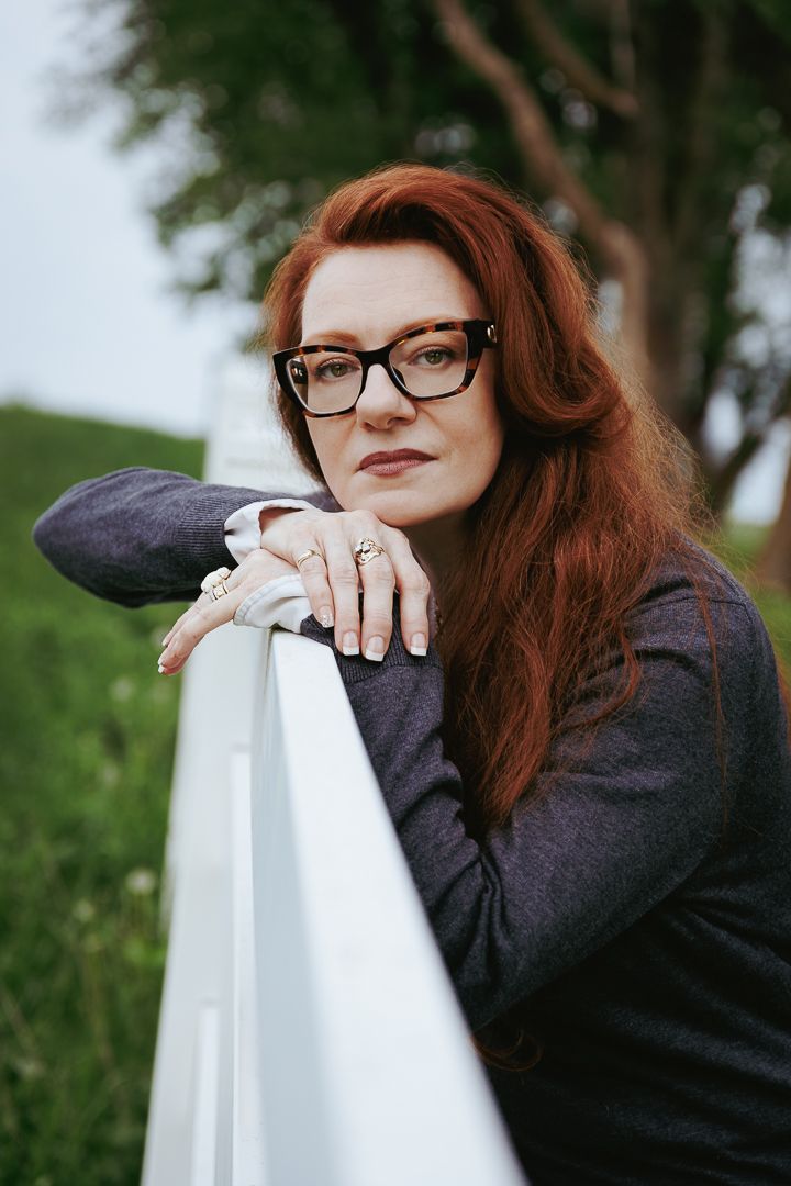 Close-up portrait of someone with red hair and glasses leaning on a white railing outdoors.