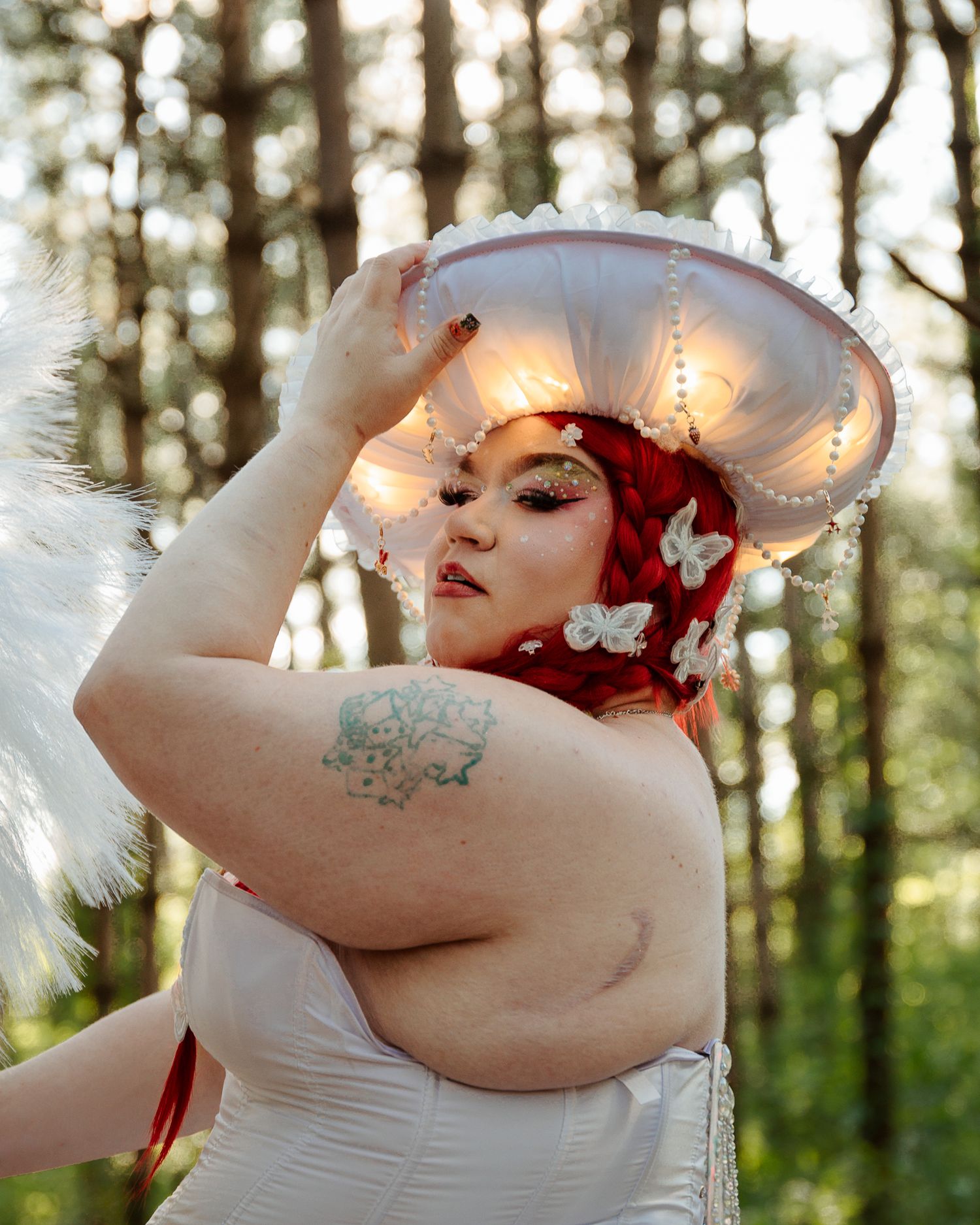 Woman with red hair and butterfly accessories wears a glowing white mushroom hat and white corset in a forest setting.