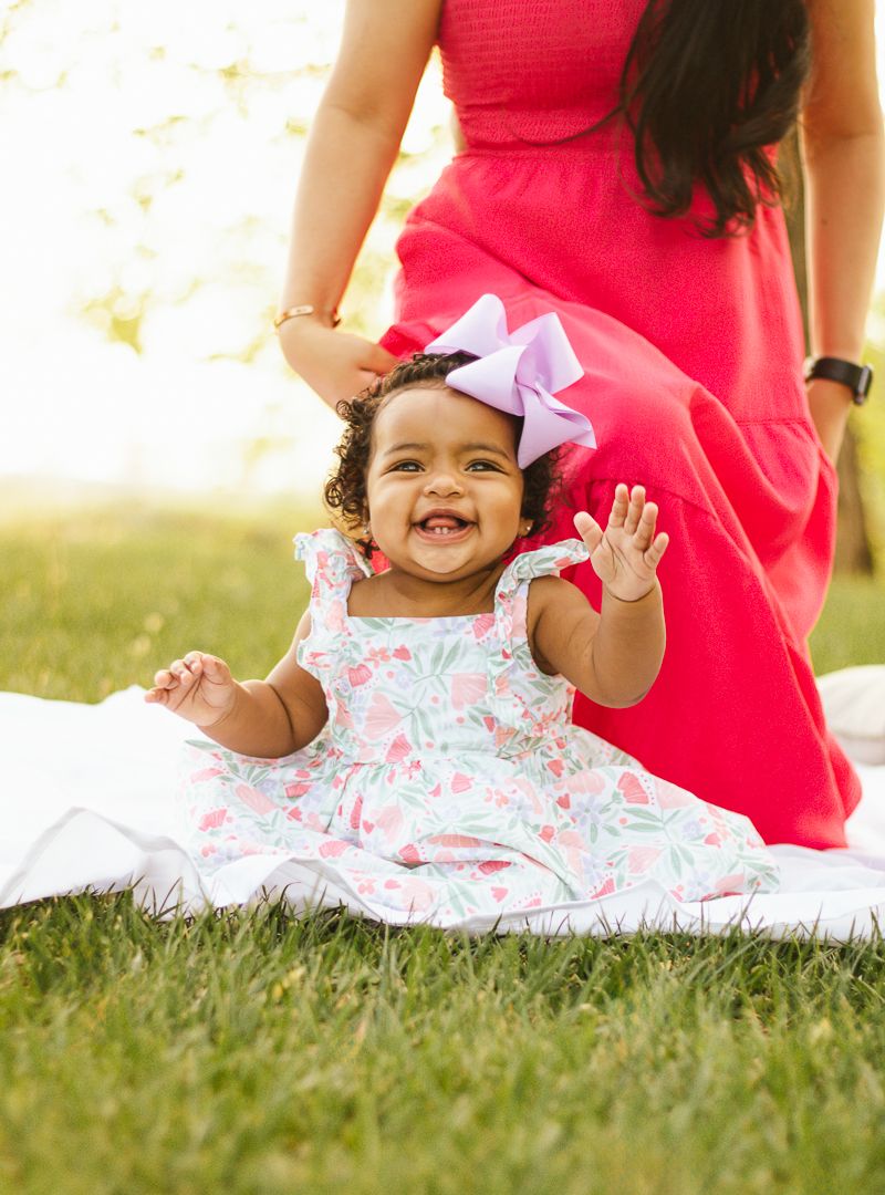 A laughing child in a floral dress plays on grass during an outdoor photo session.
