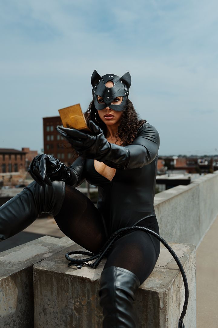 Dramatic cosplay photo of person in black catsuit and mask crouching on rooftop ledge against city skyline.