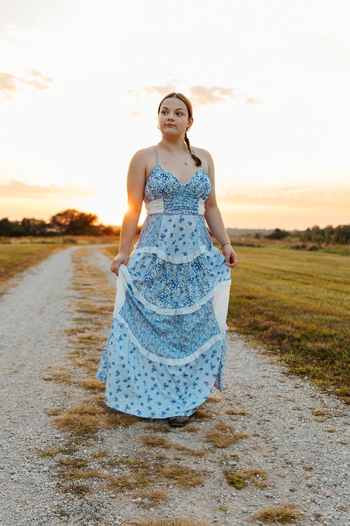 A graceful figure in a floral blue dress twirls on a dirt path during golden sunset hour.