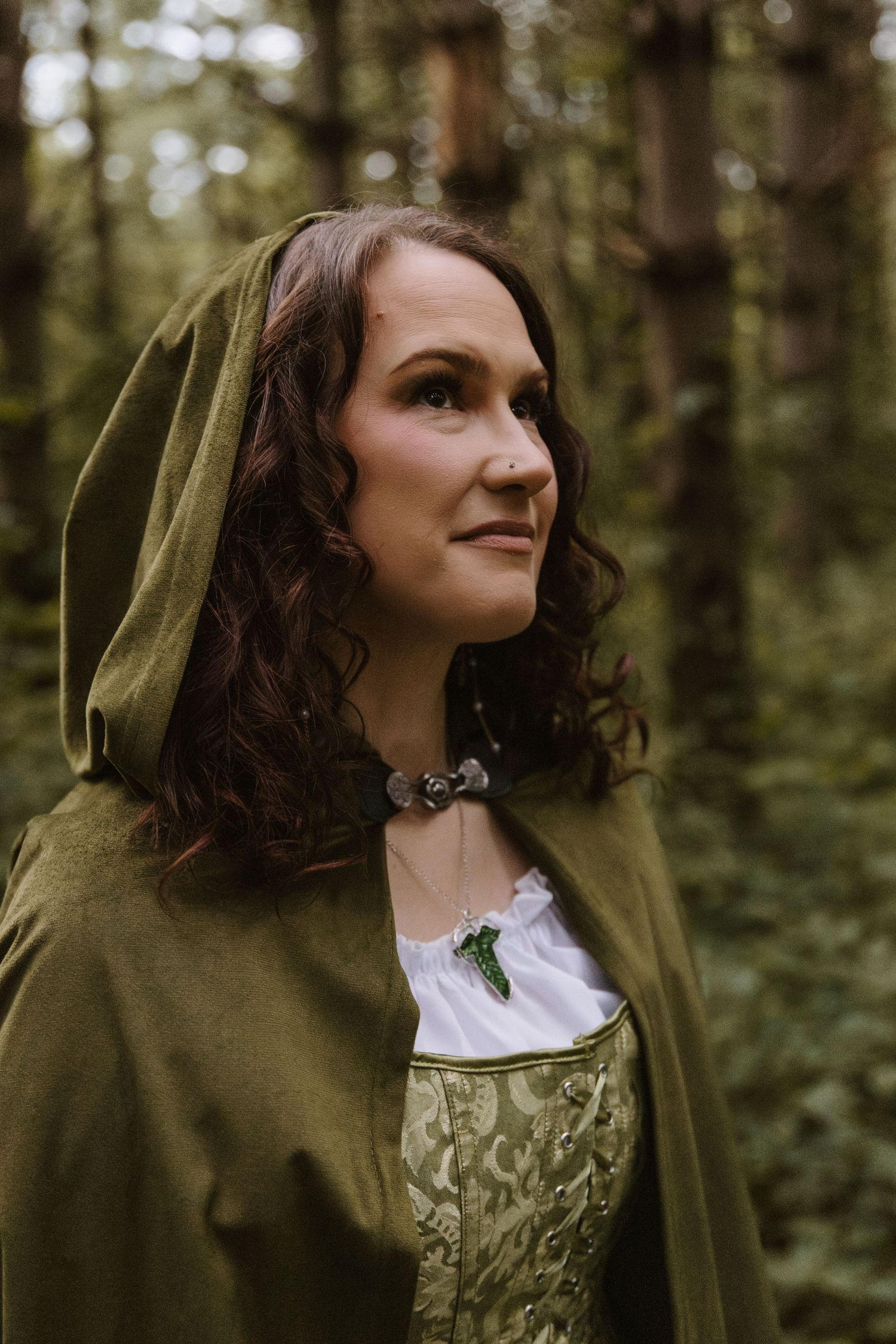 Woman in green medieval hooded cloak and corset dress poses in forest setting for Renaissance faire costume photography.
