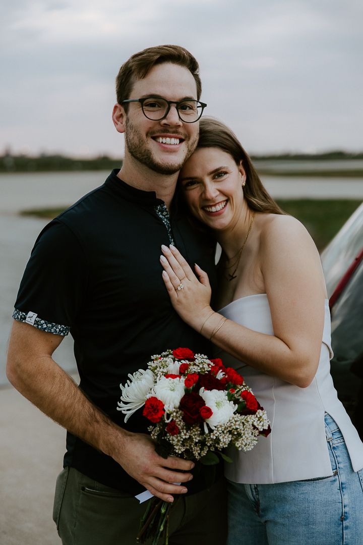Joyful couple shares intimate moment with red and white bouquet by water during sunset.