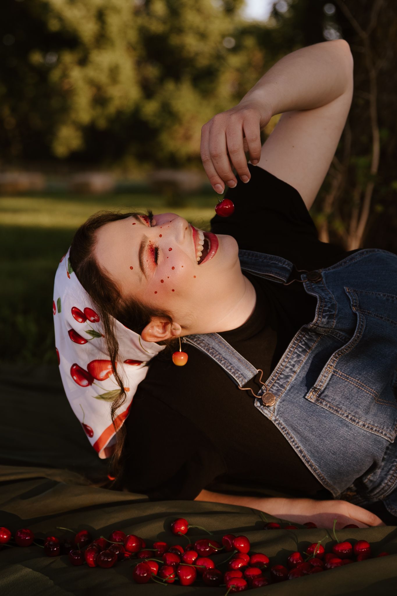Laughing woman with cherry earrings and face gems lies in grass holding cherries at golden hour.