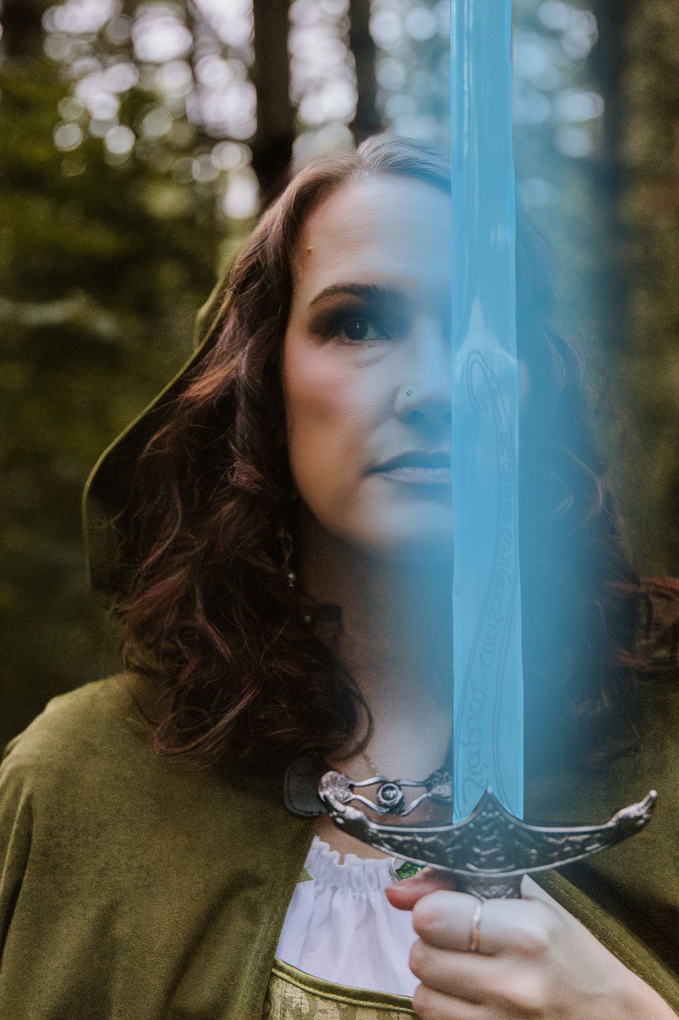 Woman in green medieval dress holds glowing blue fantasy sword in a misty forest setting.