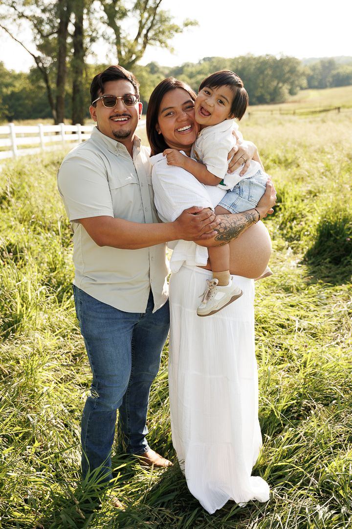 A tender family moment captured outdoors in a sunlit field with white fence and tall grass in the background.