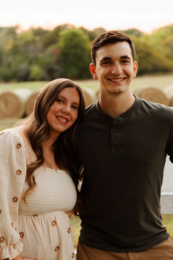 A couple poses together outdoors at sunset with hay bales in the background during an autumn photo session.