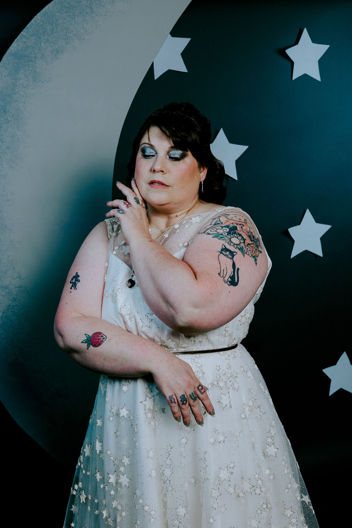 Tattooed plus-size bride posing in a star-print white gown before a crescent moon and stars backdrop.