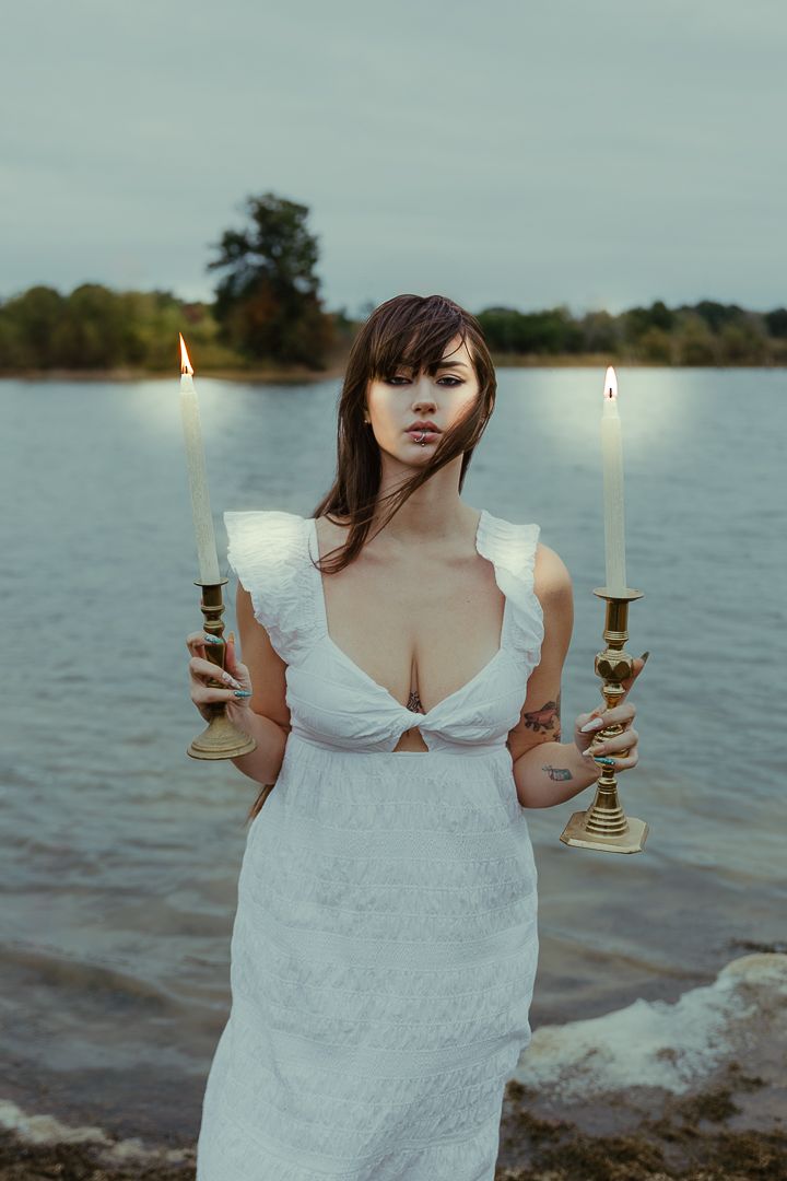 Person in white dress holds candlestick by lake at dusk in ethereal outdoor photoshoot.