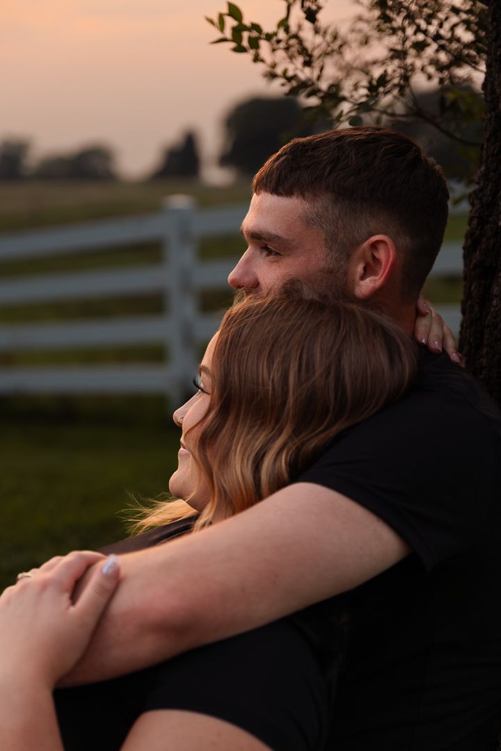 Tender outdoor portrait of couple cuddling during warm sunset light with white fence in background.