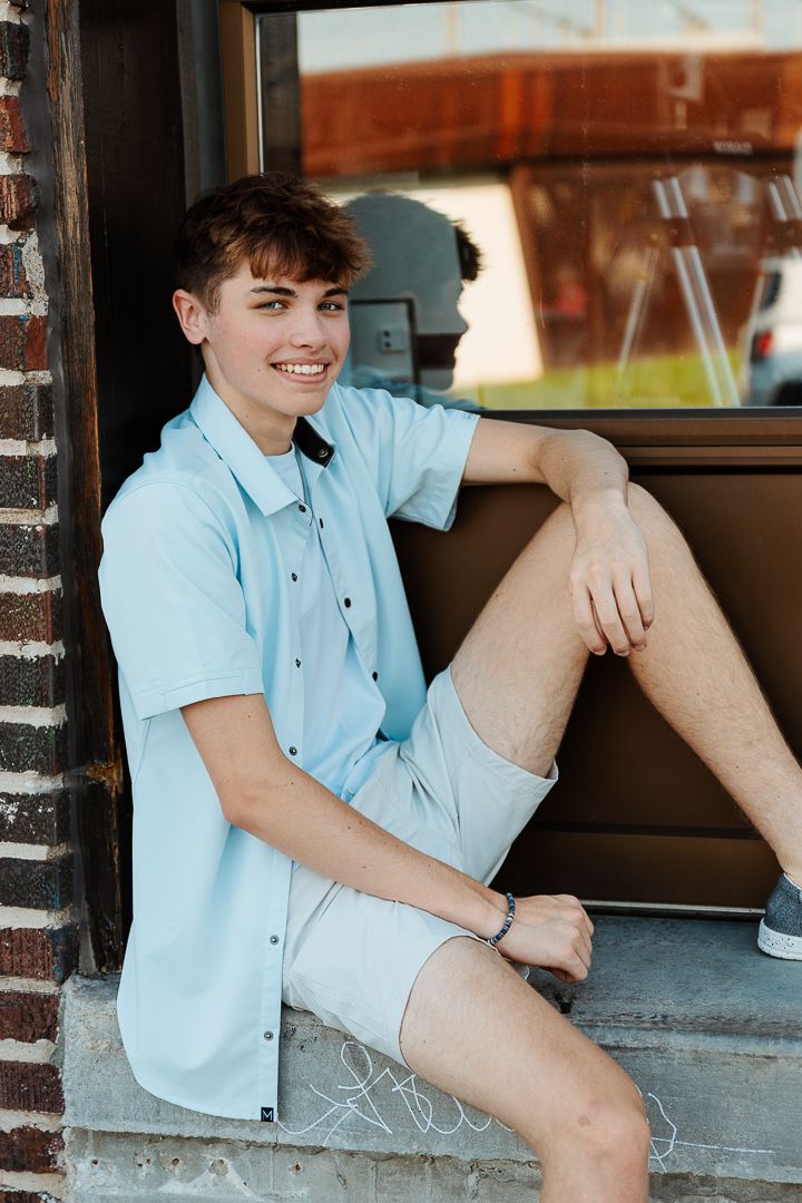A person in a light blue button-down shirt and shorts sitting by a window in natural lighting.