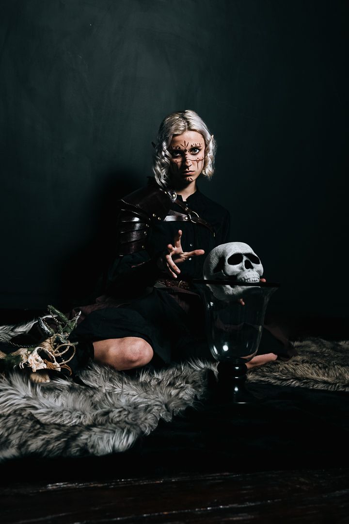 Woman with dark fantasy makeup sits on fur with skull prop in dramatic Halloween witch costume photoshoot.