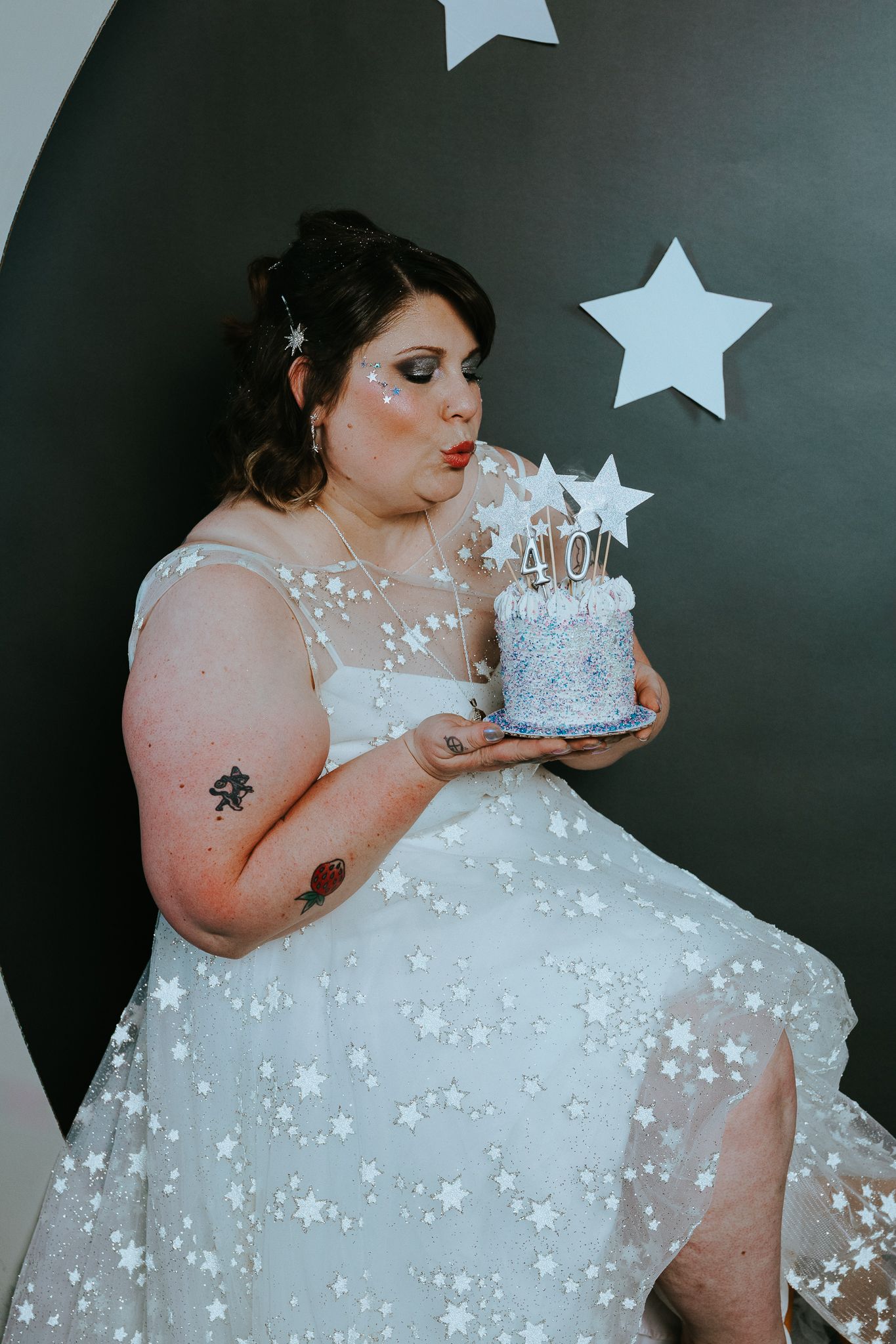 Woman in white star-patterned dress blowing candles on a 40th birthday cake, seated against a dark wall with star decorations.