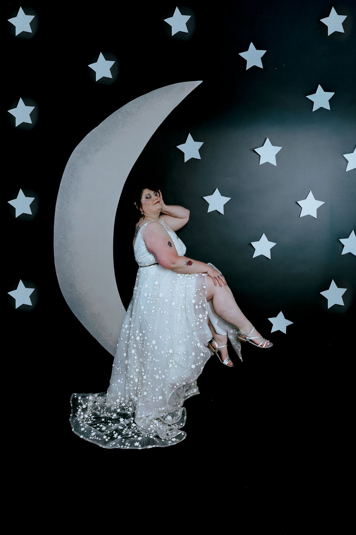 Plus-size bride in sparkly white gown poses on crescent moon prop with star-filled black backdrop.
