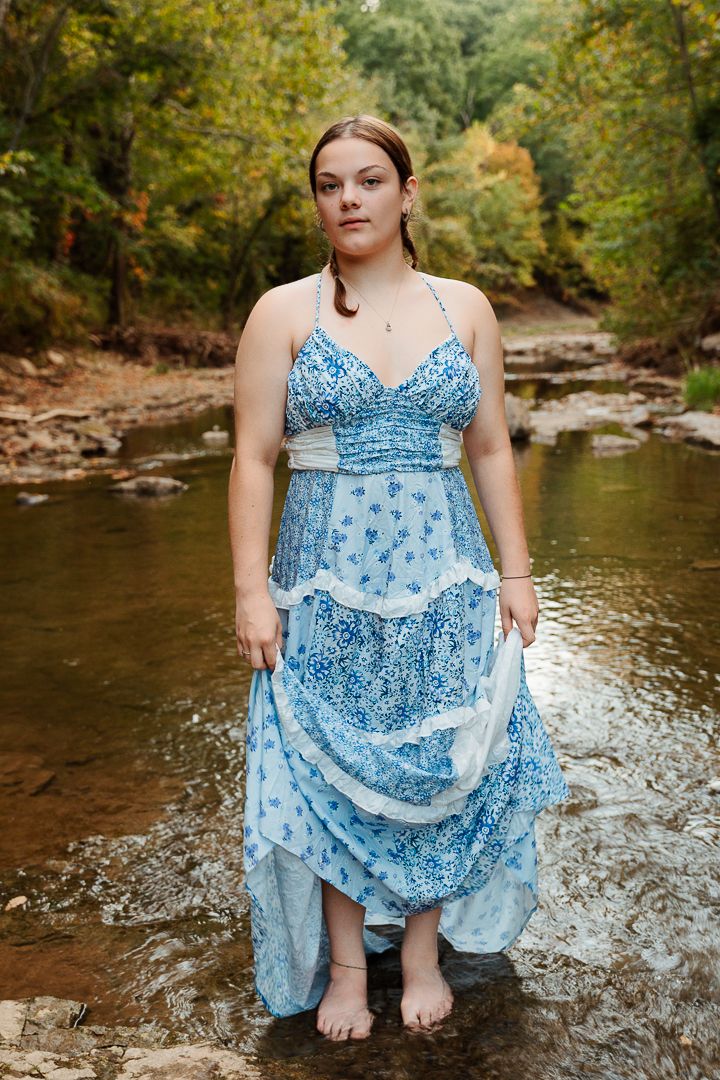 Person in a light blue patterned dress sits in a flowing creek surrounded by autumn foliage and rocks.