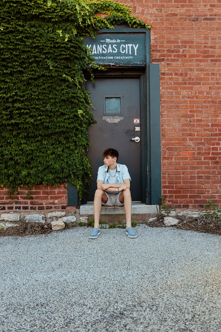 A person sits on gravel ground against a red brick wall with ivy and a dark door labeled 'Kansas City'.